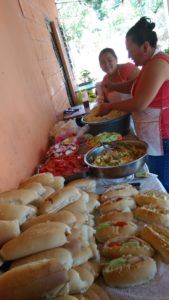 A photo of two women in aprons setting out a large table filled with sandwhiches, salad, and veggies.