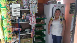 Carmen standing in her small shop with an apron on. On the shelves are various candies, chips, onions, gum, and other food.