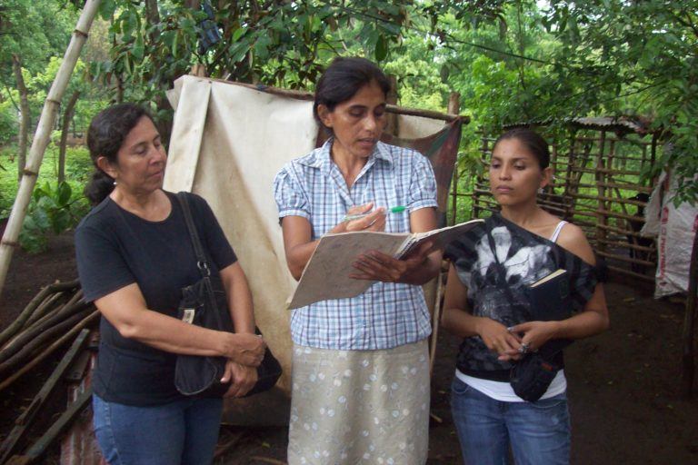 Photograph of a woman holding up a pen and reading from a notebook while two other women look on and listen.