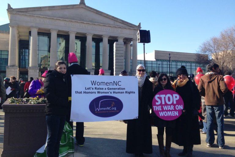 Photograph of four young women participants of Women NC in front fo the capitol holding signs that say womenNC and Stop the War on Women.