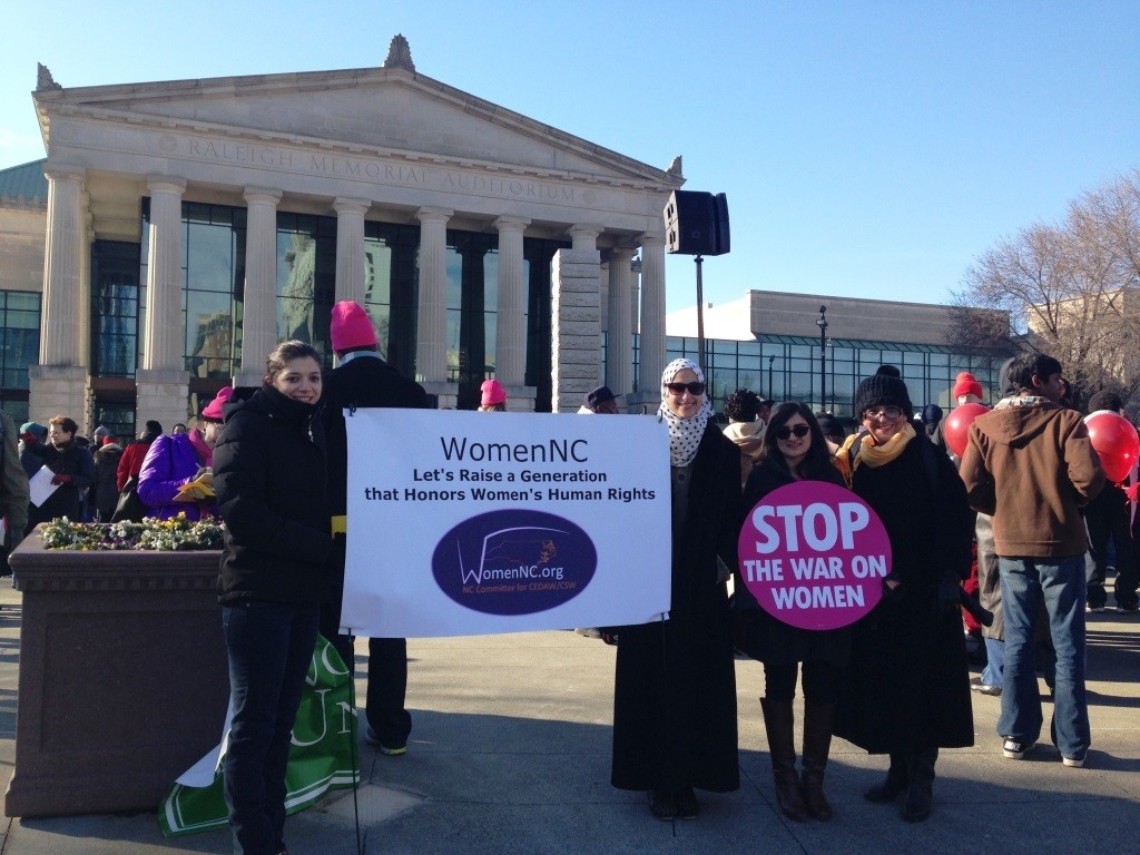 Photograph of four young women participants of Women NC in front fo the capitol holding signs that say womenNC and Stop the War on Women.