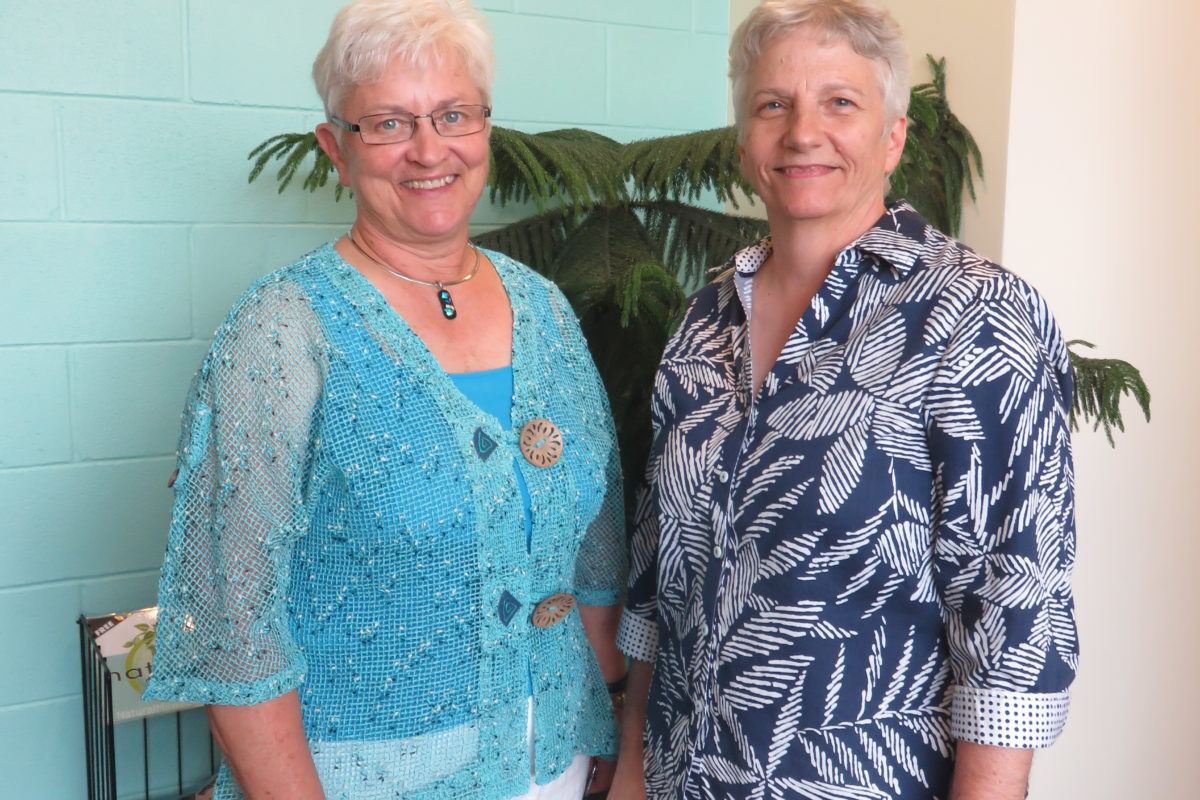 Photograph of two women in their sixties in bright tops and white pants.