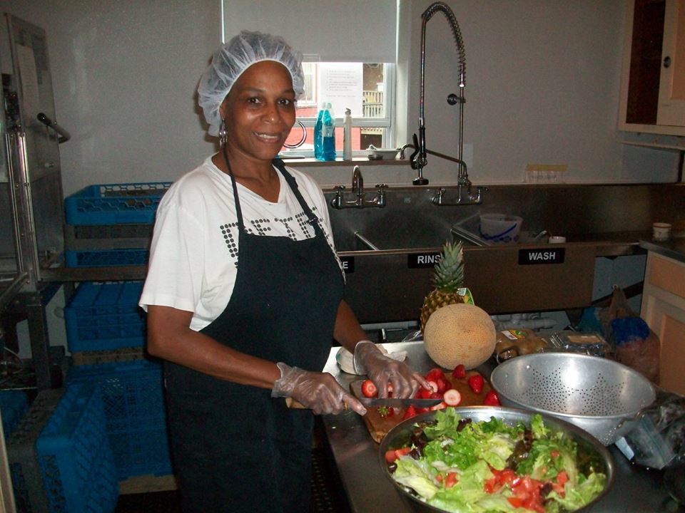 A woman with hairnet, aprons, and gloves chopping vegetables for a