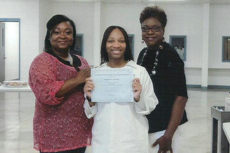 Photograph of three women, the one in the middle is dressed in a prison uniform and holding up a diploma.