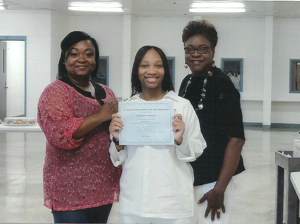 Photograph of three women, the one in the middle is dressed in a prison uniform and holding up a diploma.