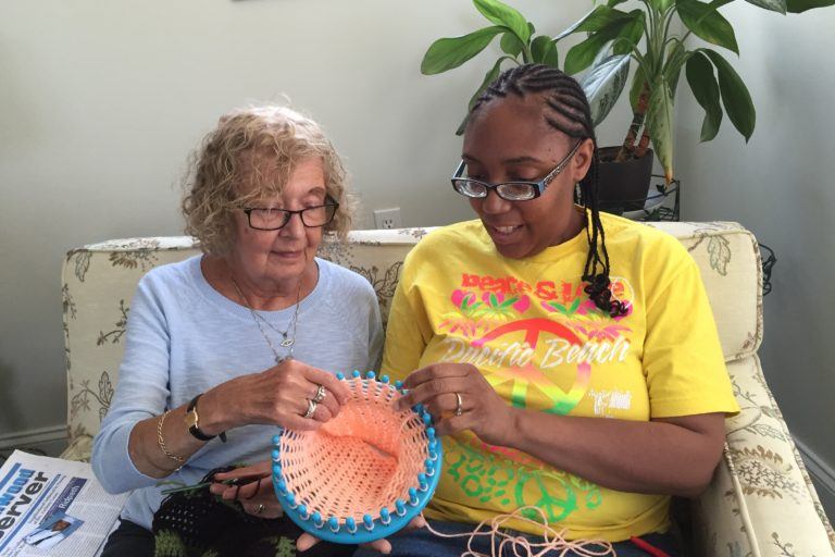 Photograph of two women in glasses sitting on a couch and making a basket together.