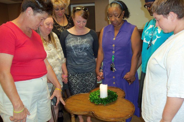 Photograph of seven women standing with their eyes closed and heads bowed around a small table with a candle on it.