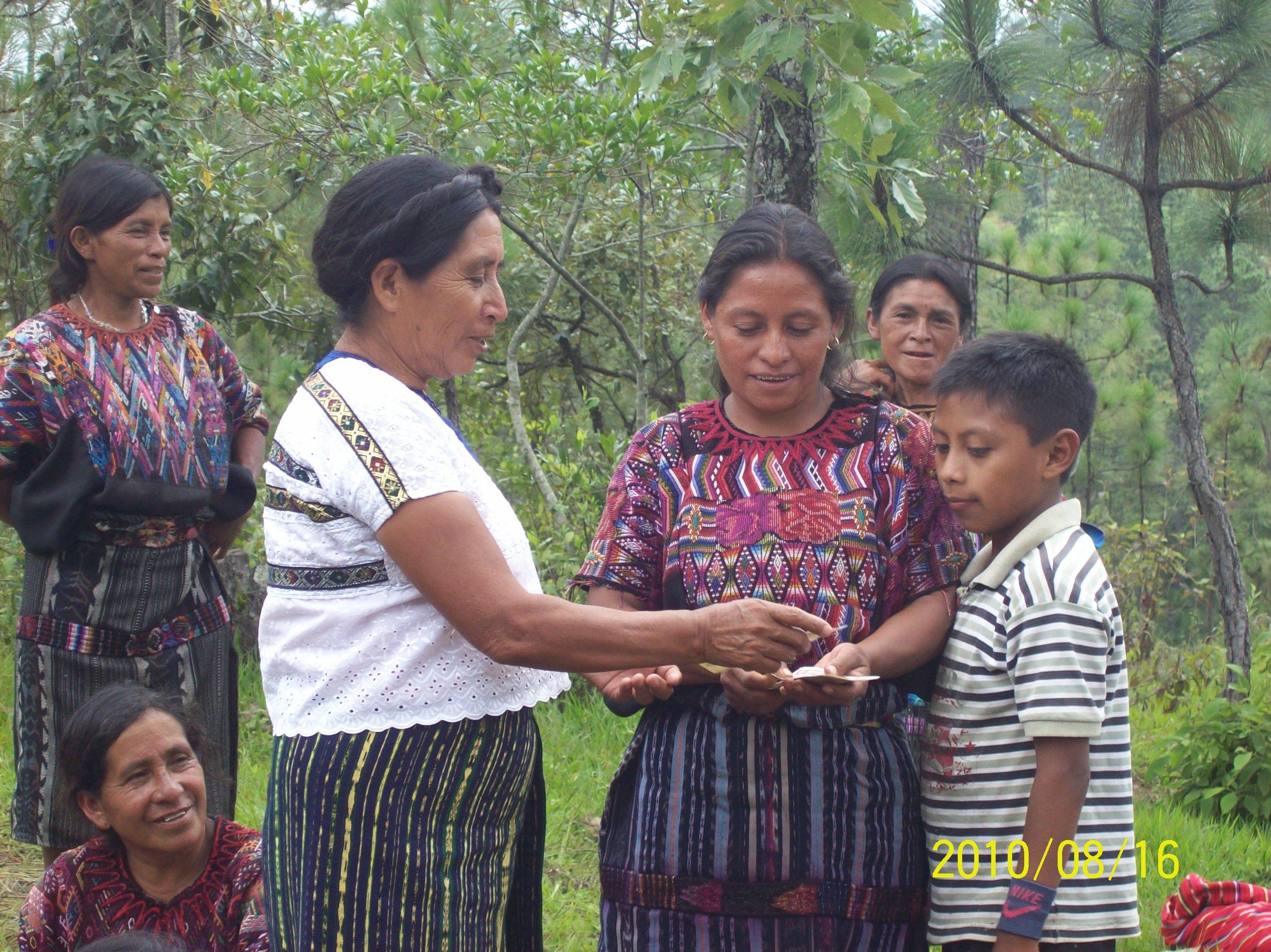 Photograph of a woman handing another woman a loan while her son looks on.