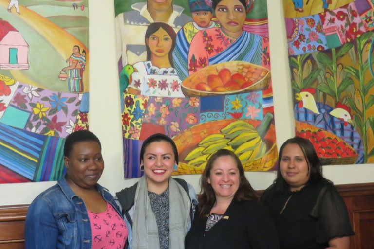 Photograph of four women with a bright wall mural of a mother and daughter holding a tray of food behind them.