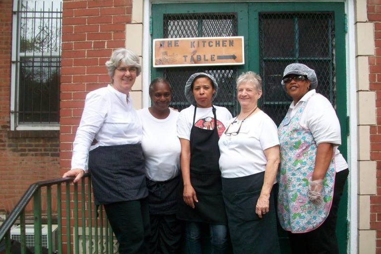 Photograph of five women on the steps in front of a church with aprons and hairnets on. There is a sign behind them that says The Kitchen Table.