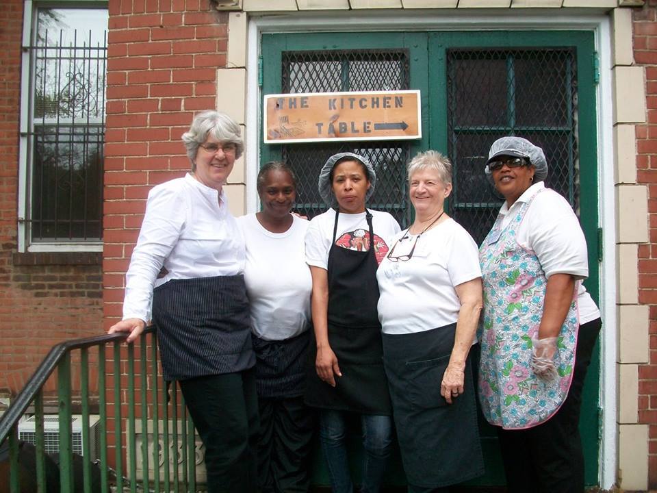 Photograph of five women on the steps in front of a church with aprons and hairnets on. There is a sign behind them that says The Kitchen Table.