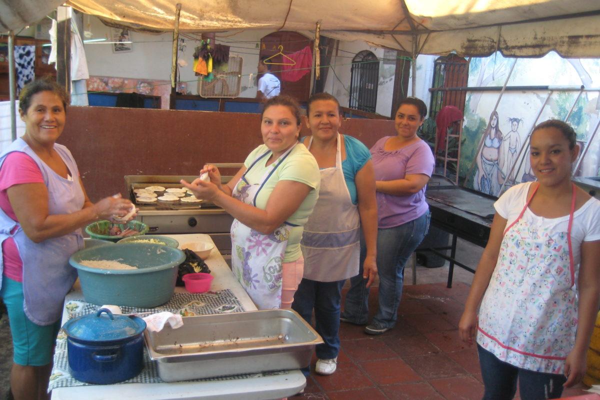 Women with aprons on all in different stages of making tortillas. Some are mixing dough, some are putting them on a grill.