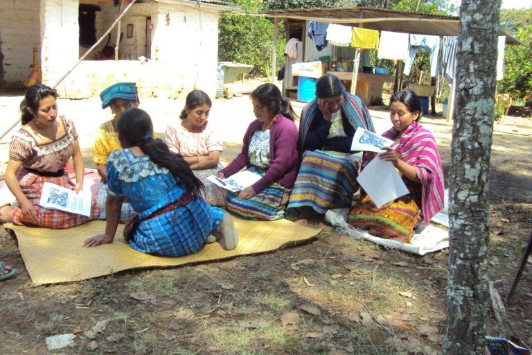 Photograph of a women in traditional guatemalan dress sitting on the ground on mats writing on papers.