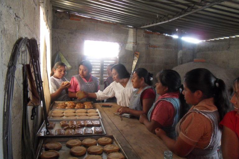 Photograph of several women in aprons around a high counter with baked bread on it.