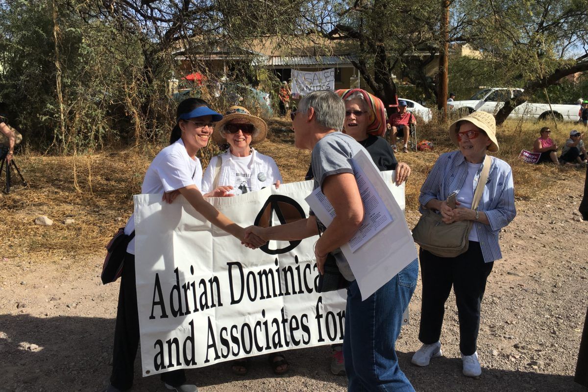 A woman shakes hands with another woman holding up a sign that says Adrian Dominicans and Associates for Peace