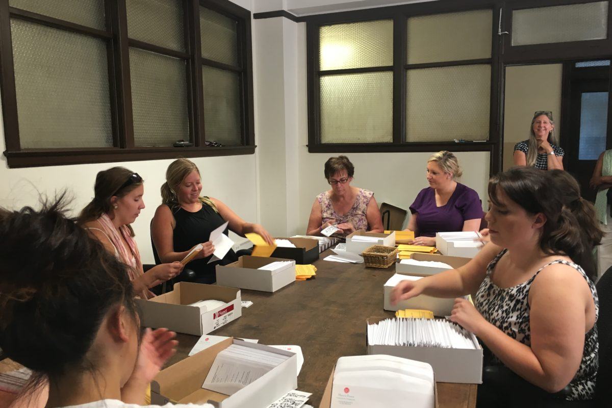 A group of young women sit around a table stuffing envelopes and chatting.