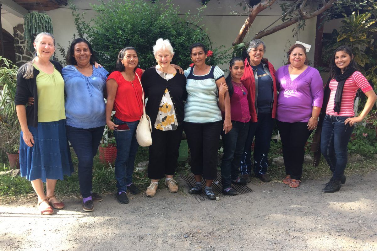 A group of eight women, two white, the rest latina, stand with their arms around one another smiling.