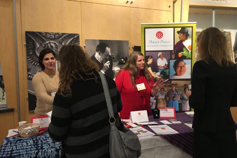 Two women stand behind the Mary's Pence exhibit table, smiling and talking to two women in front of the table.