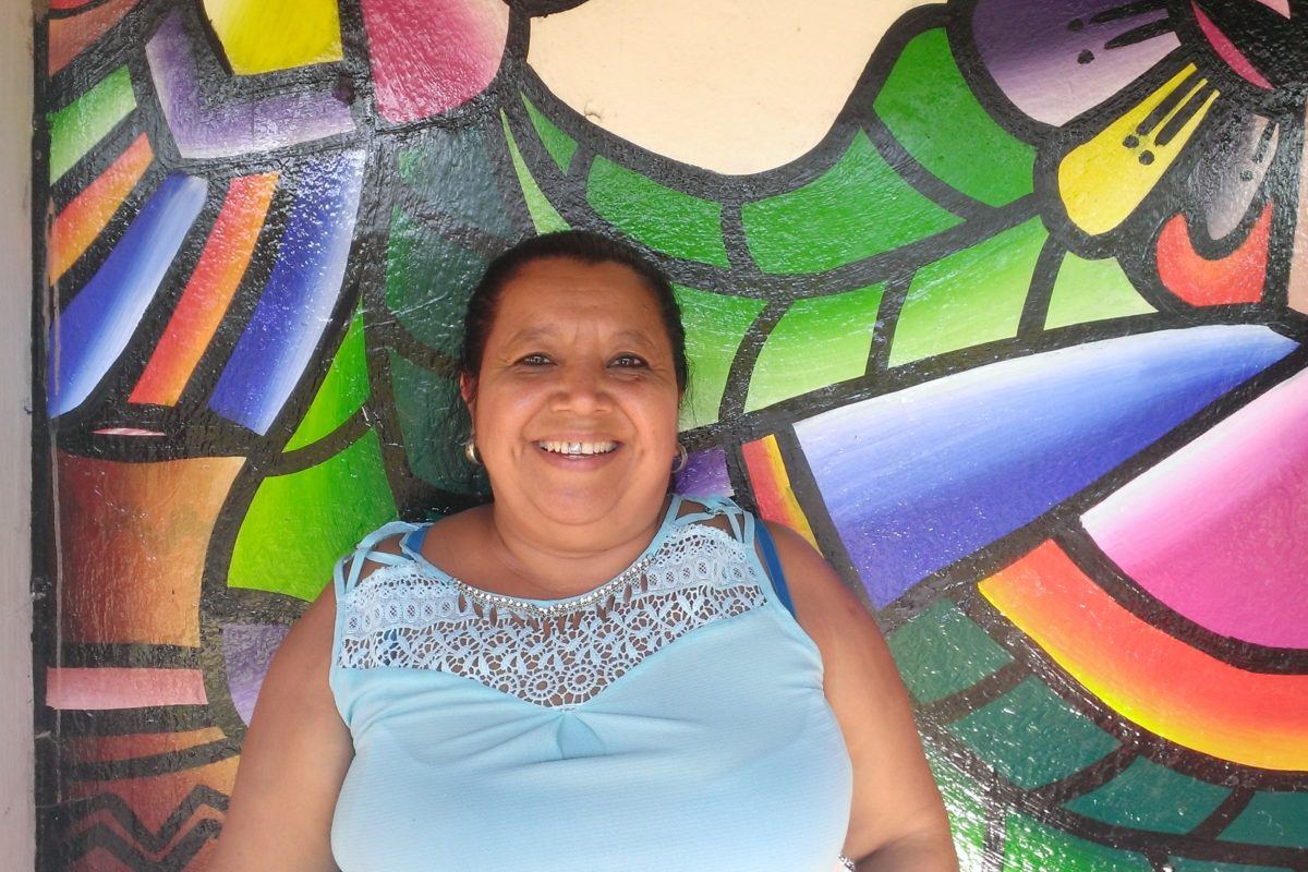 A middle aged woman smiles broadly, she is standing in front of a wall painted in bright colors in the traditional salvadoran style.