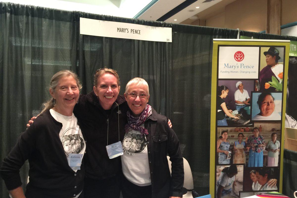 Photograph of three woman standing at a Mary's Pence exhibit booth. Two women are wearing Mary's Pence t-shirts.