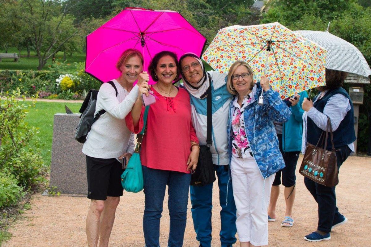 Four women - 2 iraqi, 2 white americans - smile with their arms around one another. The women on the ends hold umbrella's over the group.