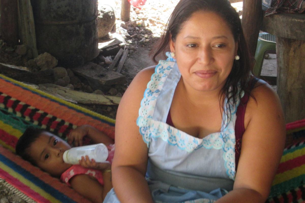 Salvadoran woman wearing a light blue apron sits on the edge of a hammock. Behind her in the hammock a toddler lays drinking a bottle.