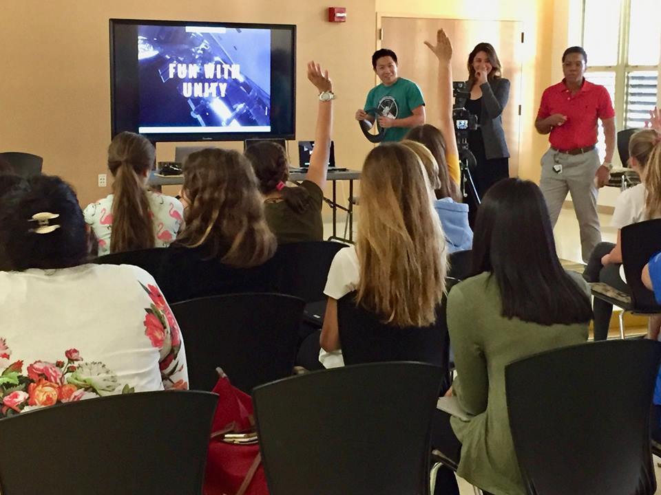 Photograph of a classroom of young girls from the back, the teacher stands up front and two girls are raising their hands.