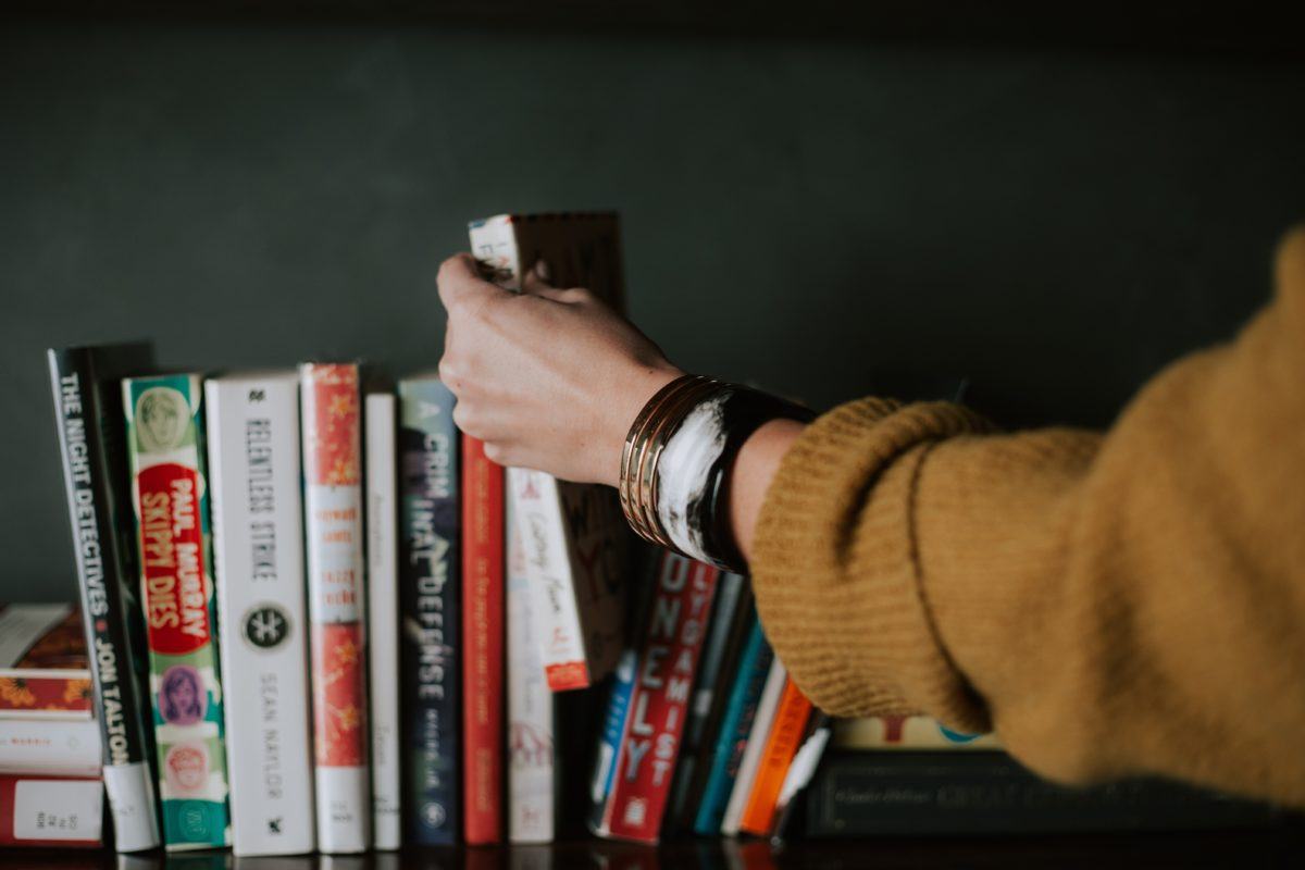 Photograph of a woman's arm and hand picking up a book from a shelf full of different colored books.