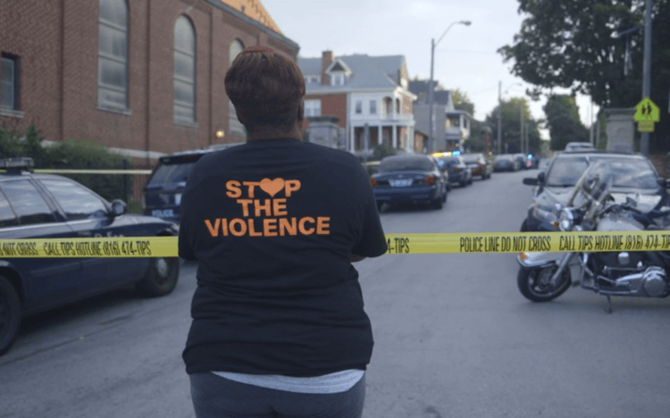 Photograph of a woman from the back, looking across police tape. Her shirt reads Stop the Violence.