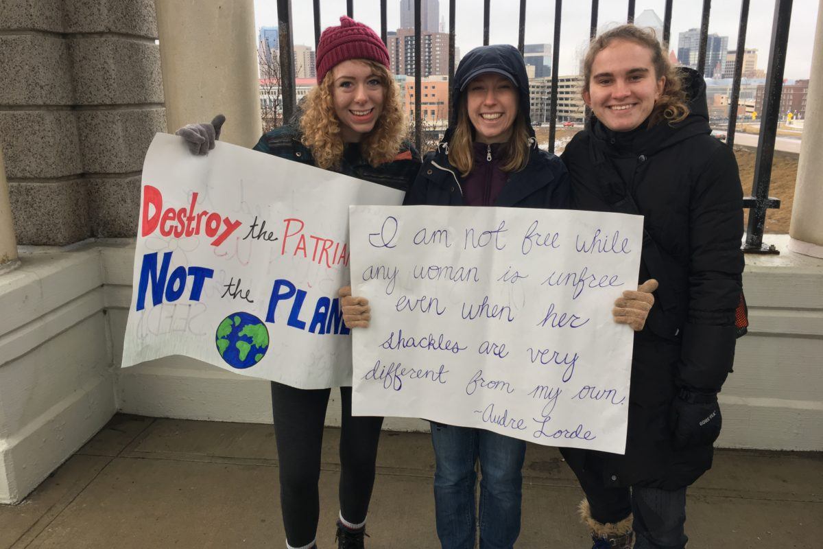 Photograph of three young women holding signs that read "destroy the patriarchy, not the planet" and "I am not free while any woman is unfree, even if her shackles are very different than my own.. - Audre Lorde"
